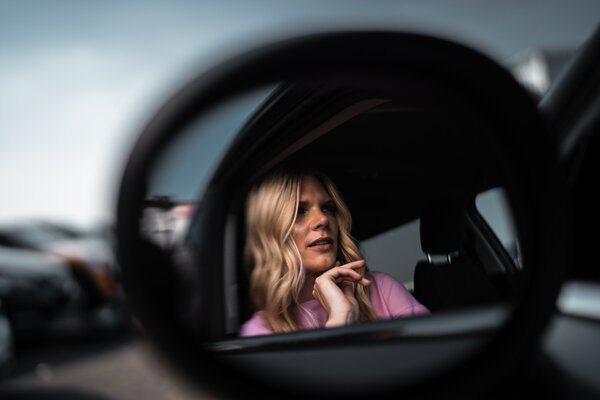 Support beim Abschließen eines Auto Abos Die Spiegelung einer im Auto sitzenden Frau im Seitenspiegel, die fragend die Finger unter dem Kinn hält.